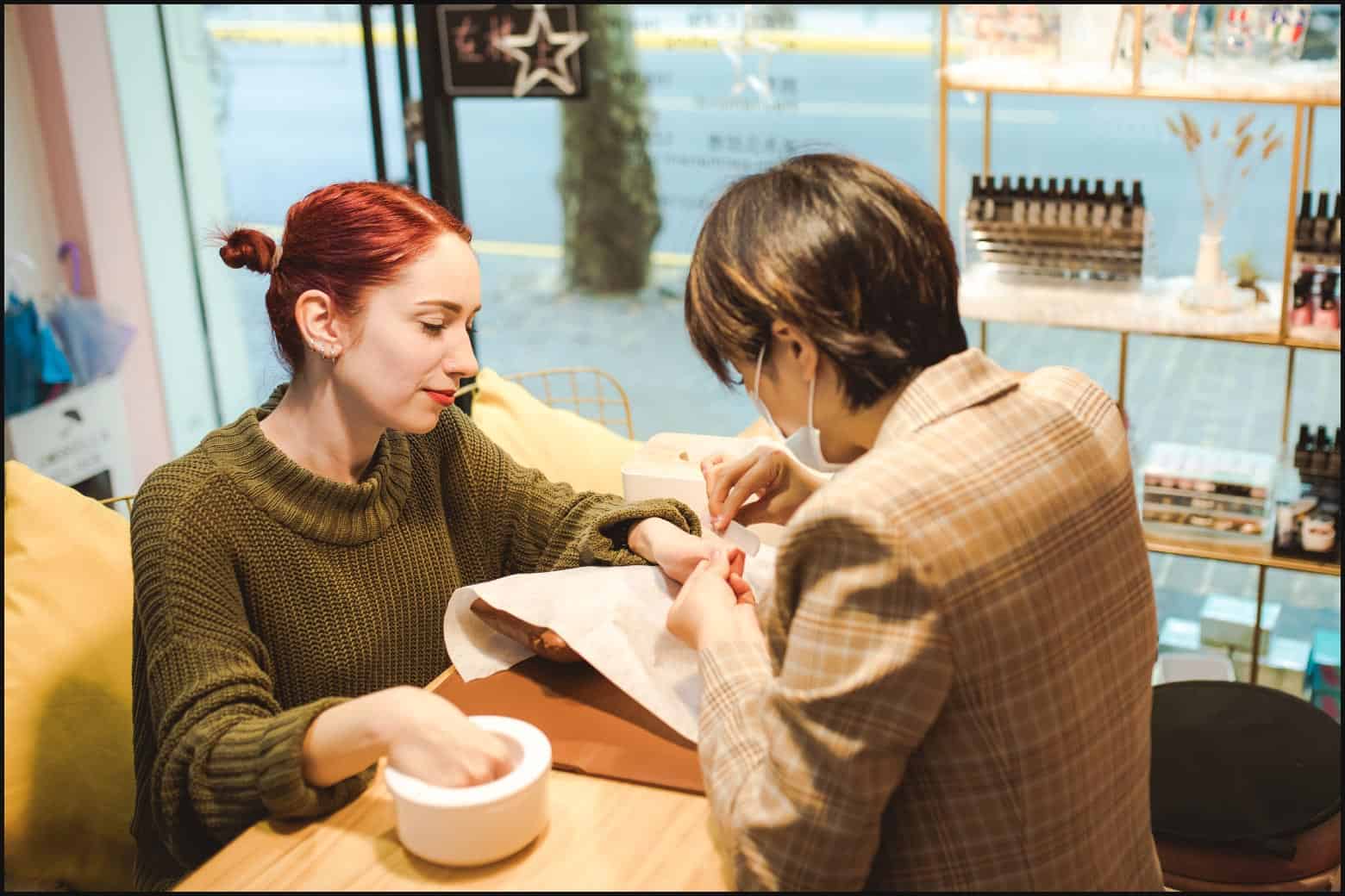 client on left side of table, wearing glasses and pink sweater, getting nails done by nail tech on opposite side